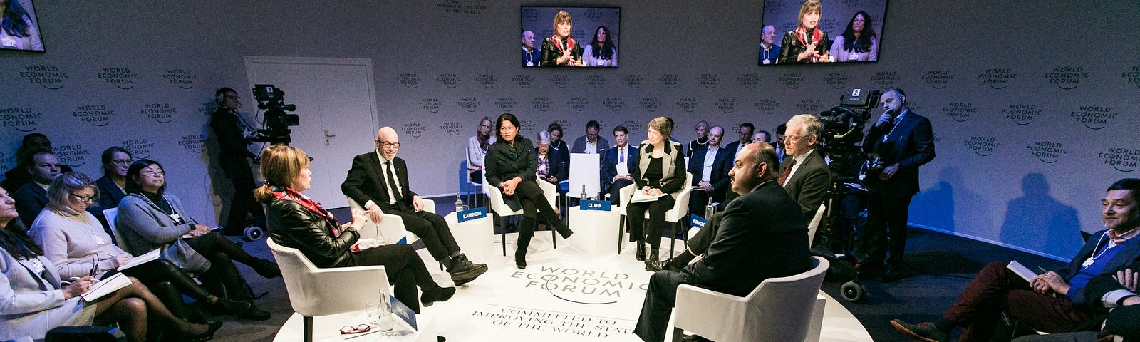 A group of speakers sit on a round platform as they particiapte in a public forum. They are surrounded by an audience and on the wall behind them are TV’s broadcasting thier discussion.