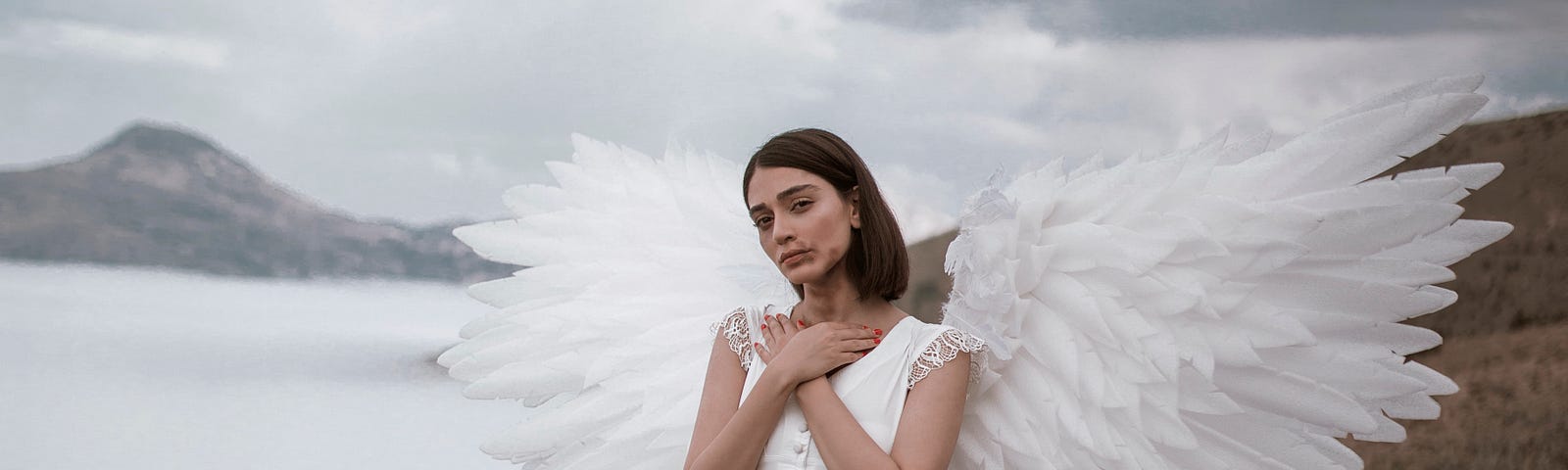 A young woman in a white dress and with large white wings, stands on a rock in front of a blue sky and water landscape, hands folded across her chest and staring innocently to the camera.