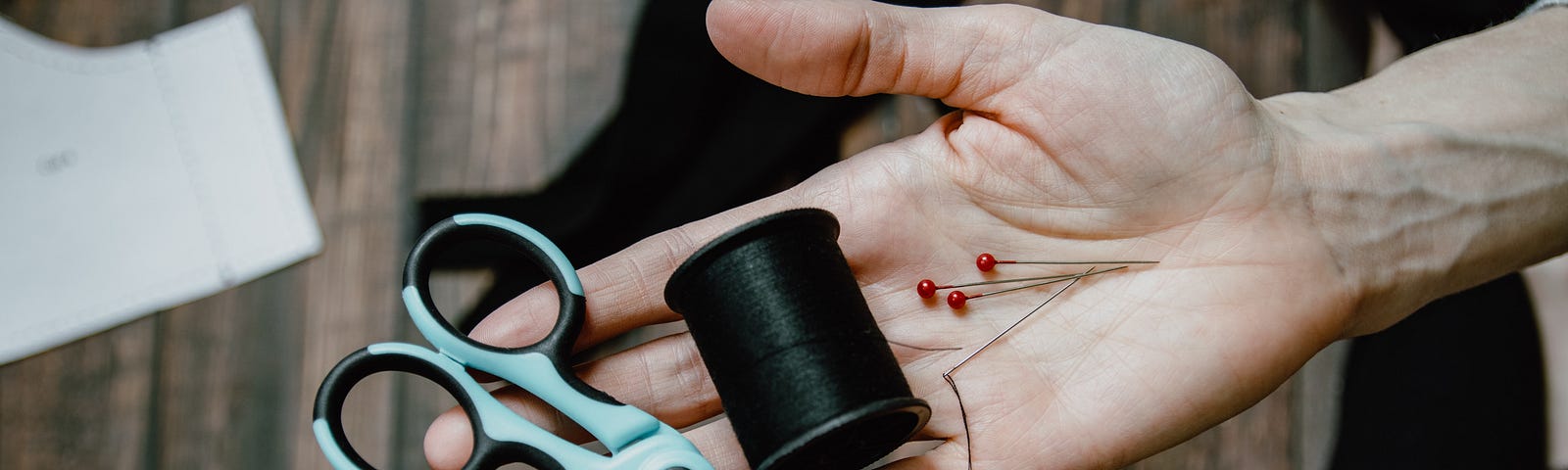 An older person’s wrinkled hand holding out scissors, thread, and pins in the palm of their hand over a work table with fabric scraps and patterns