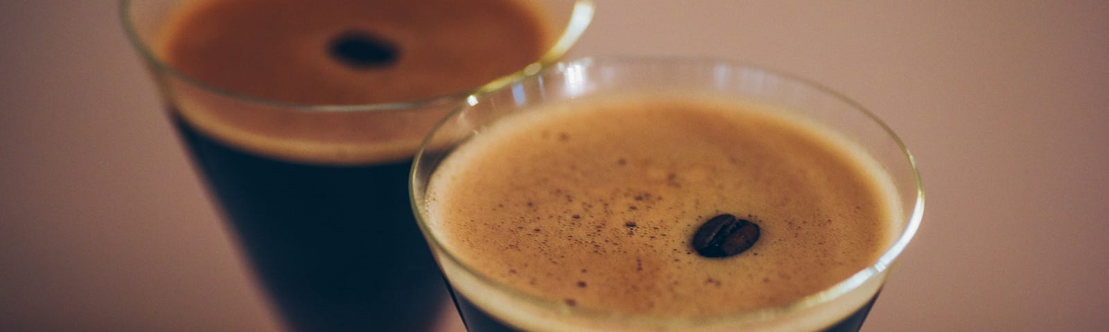 Two conical Martini glasses full of dark brown liquid with a foamy top and a coffee bean in the centre are placed on a counter top