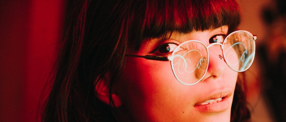 Young brunette woman with glasses thoughtfully looking at the camera in red light, with her glasses reflecting some green neon