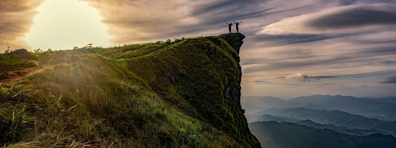 Two people on the edge of a cliff with a night sky and sunset behind them.