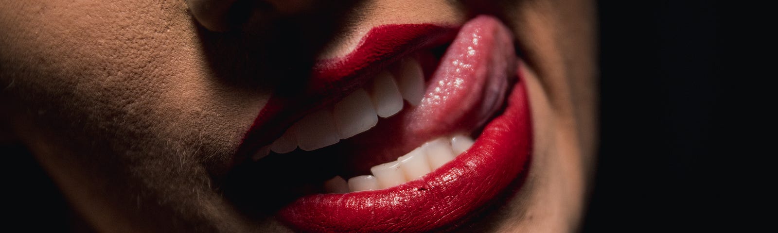 Partial image of the face of woman, red lips and her tongue sticking out the left corner of her mouth. Part of her nose is visible.