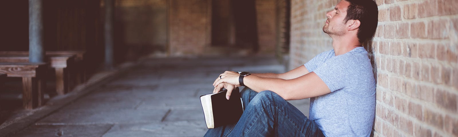 A guy sitting leaning against a wall with a book in his hand and his eyes closed as if sleeping.