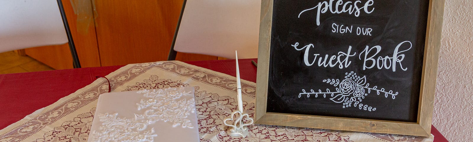 Small card table covered in pink cloth & lace with white guest book, and a chalk board sign that says please sign guest book