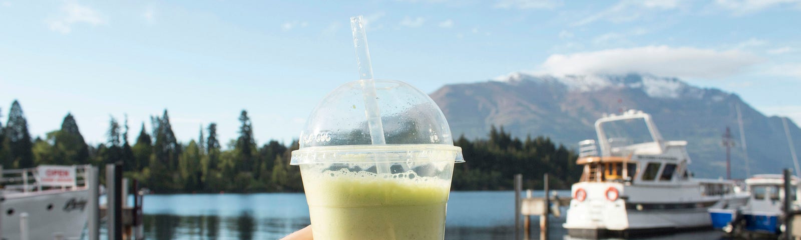 A hand holding a green smoothie in a clear cup, with a scenic background of a lake, boats, and mountains, symbolizing a refreshing and healthy lifestyle.