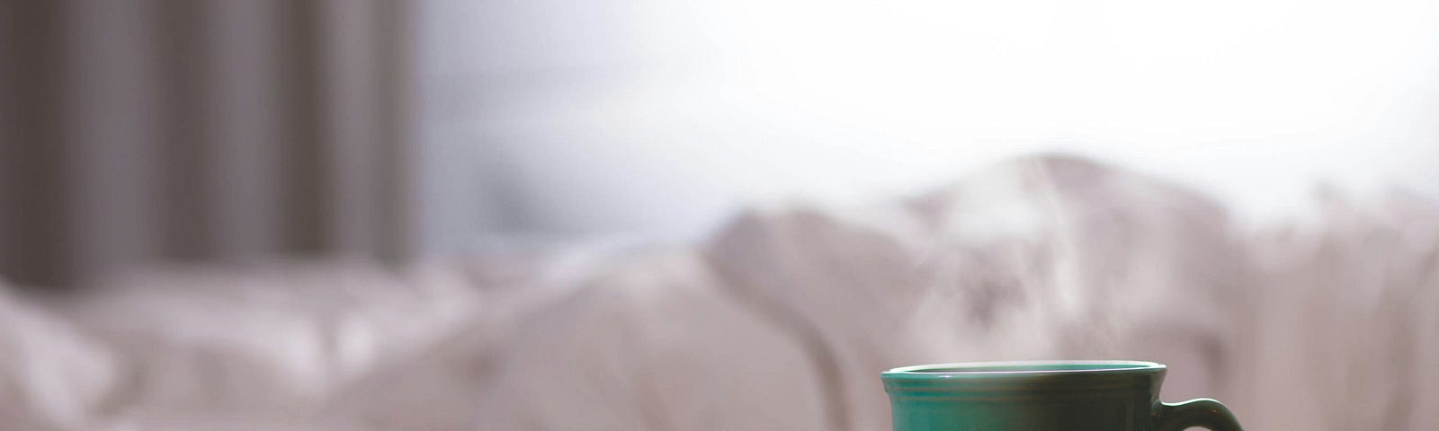A steaming green coffee cup in the foreground. A sleeping figure, wrapped in a white comforter in the background.