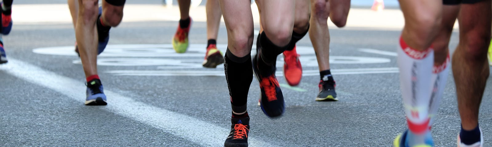A group of legs, all pictured from below the upper thigh, wearing shorts and colorful running shoes, pound the pavement in what looks like an organized running race.