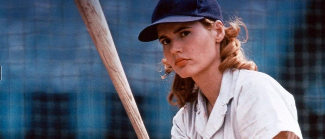 A white woman in her thirties with shoulder-length blonde hair stands at home plate. She holds a baseball bat, wears a uniform and blue ballcap, and has a determined look.