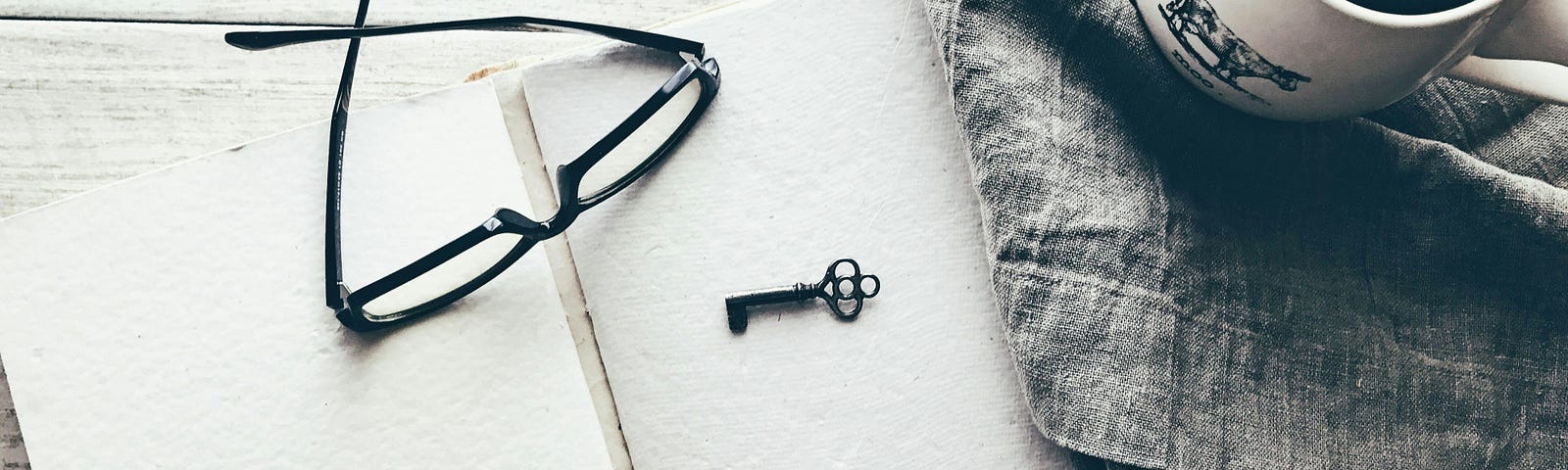 Black and white photo of a wooden table on which, from right to left, there is a cup with what looks like coffee, on a denim placemat, an open journal with white sheets, on top of it a pair of glasses and a key.