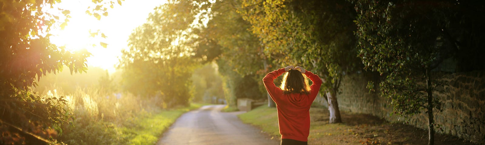 A blonde woman with an orange sweatshirt and black pants walks along a wide path, with trees on either side and sun in front of her