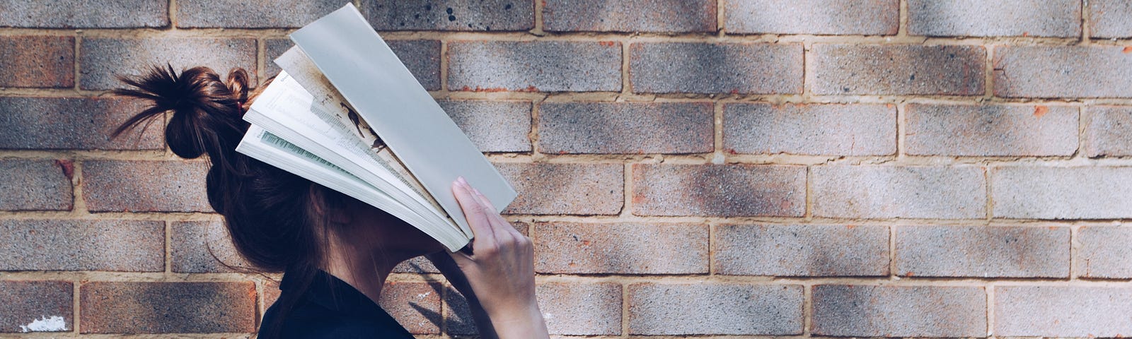 A woman stands in front of a wall and holds an open book in front of her face.