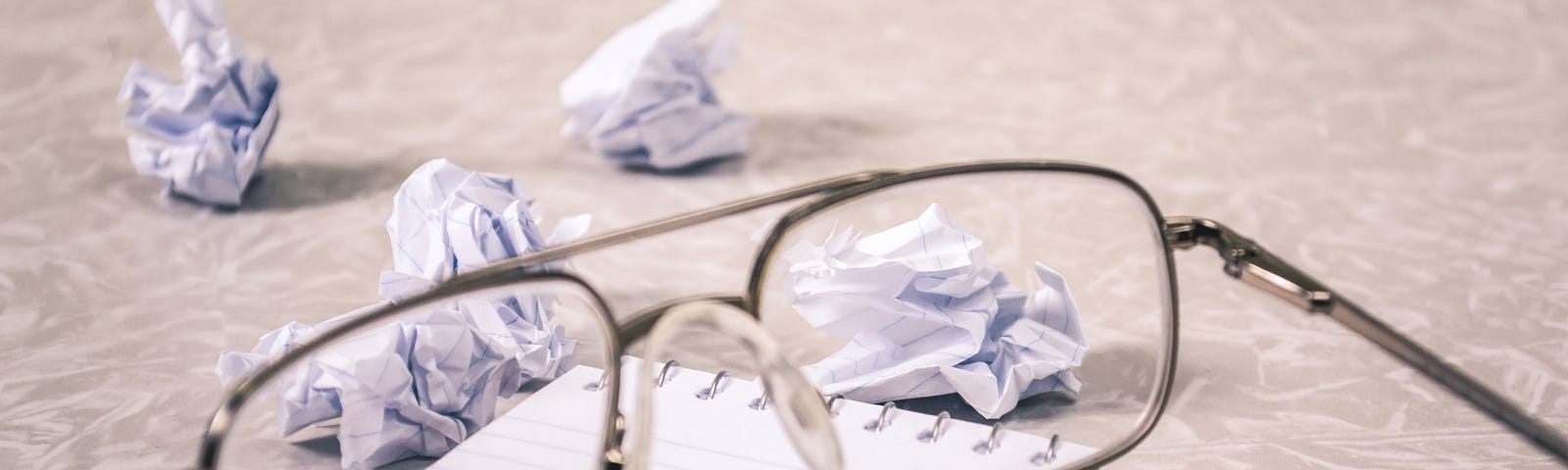 A pair of eyeglasses on top of a notepad with crumpled pieces of paper next to them.