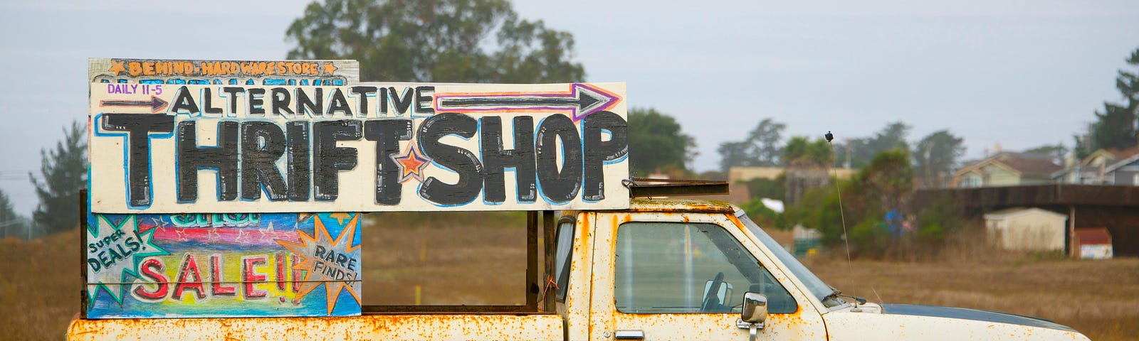 An old white truck carrying a large, hand-painted sign that says, Alternative Thrift Shop Sale.