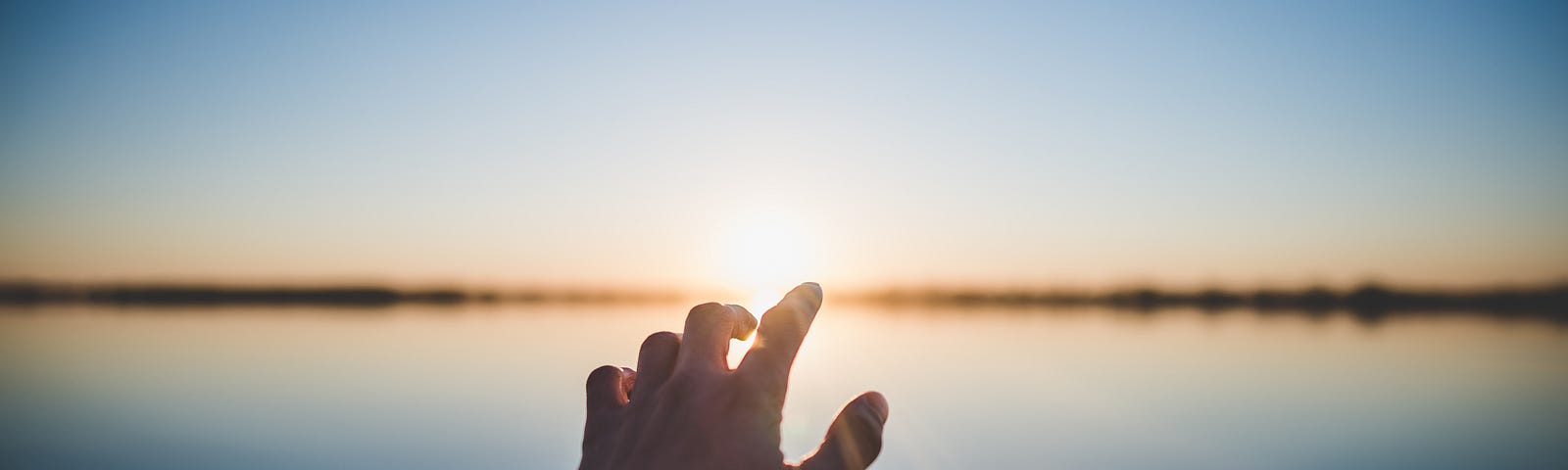 Hand reaching into the horizon, above a body of water, sun shining in the background.