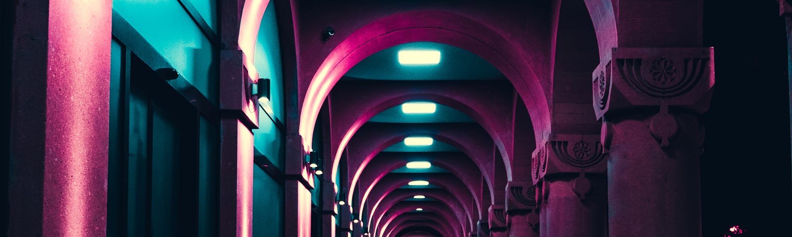 A long hallway with pink arches. Square fluorescent lights stud the ceiling between arches, and there are sconce lights on the wall on the left.