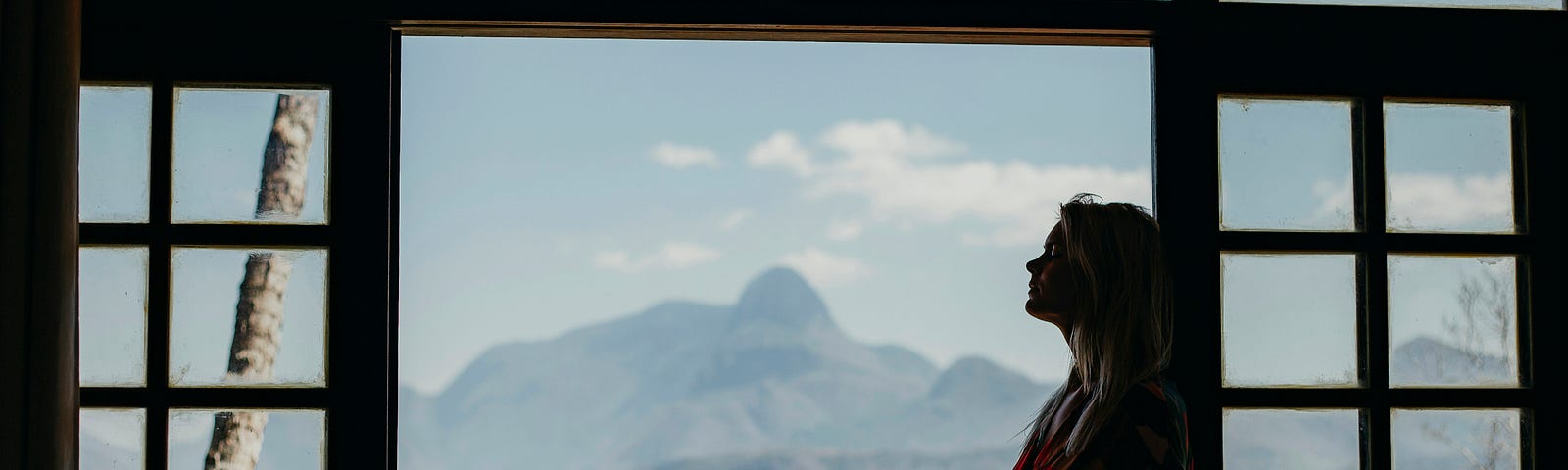 A silhouette of a person standing in front of a large window, gazing at a scenic view of mountains and a clear sky, symbolizing contemplation and the journey towards overcoming addiction.