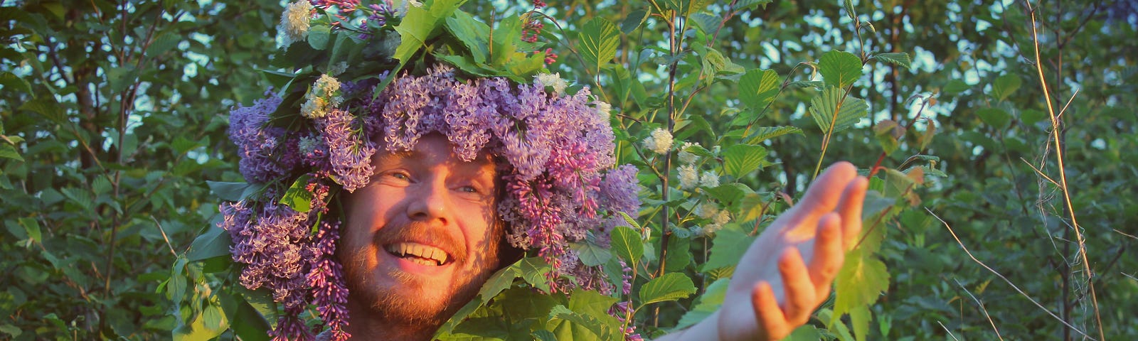 The goofiest picture of a bearded guy in a hat made of purpose blossoms and green leaves, smiling and beckoning others to join him.