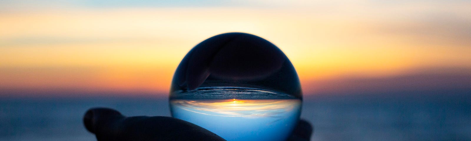 A dark hand holding a glass sphere in front of a beach. The image in the sphere is darker than the background.