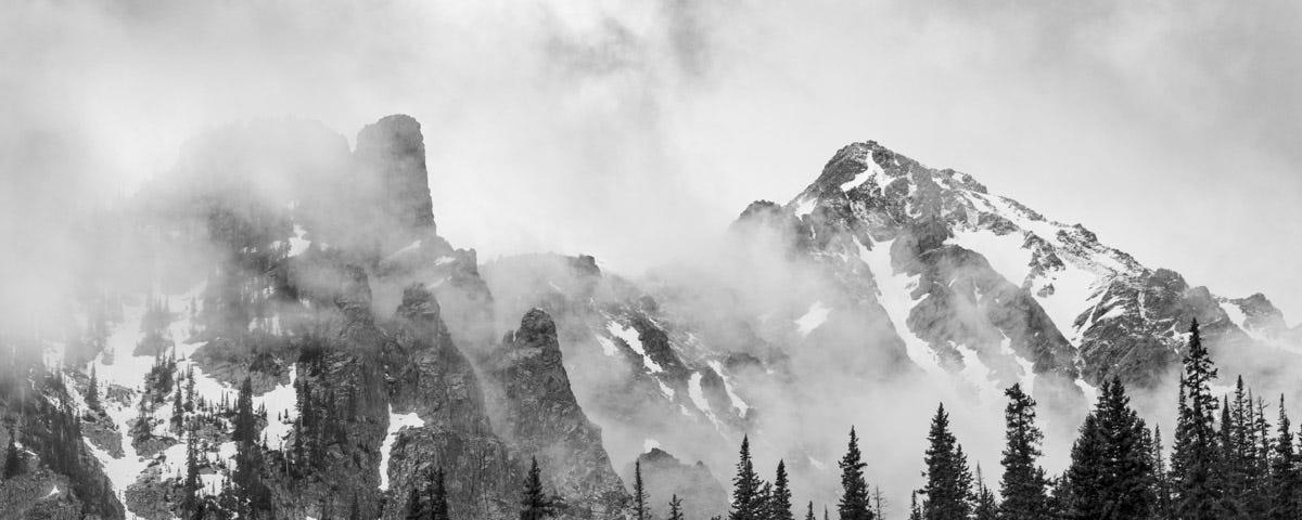 A black and white image mist lifting off rugged mountains that tower over a forest of evergreens.