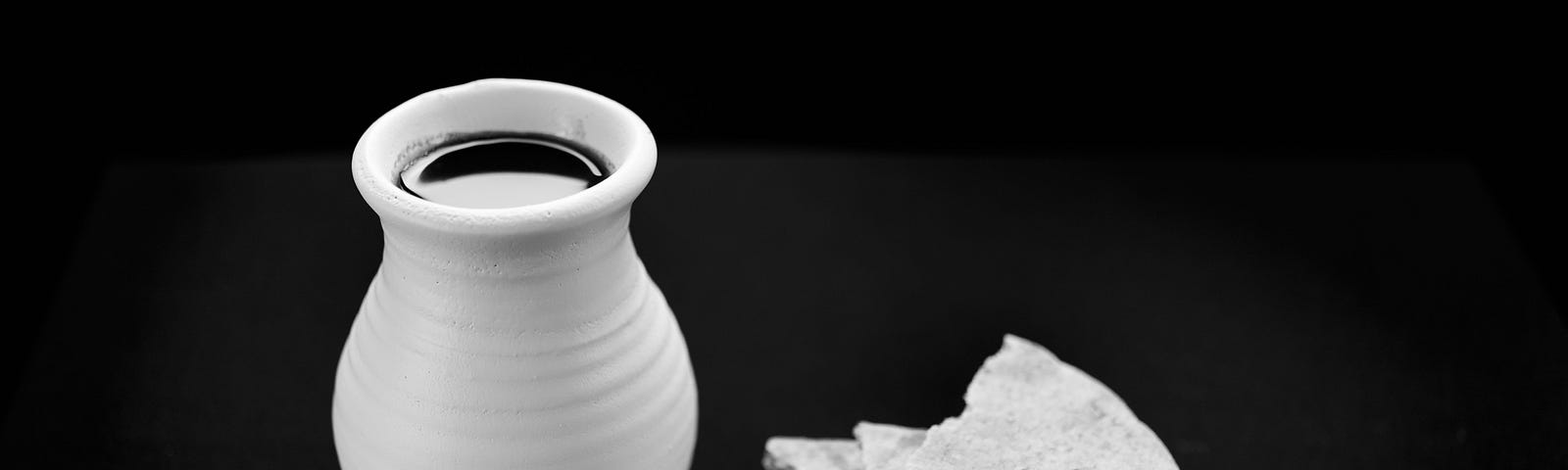 White, ceramic urn of dark wine; white ceramic plate of broken flatbread. Black background.
