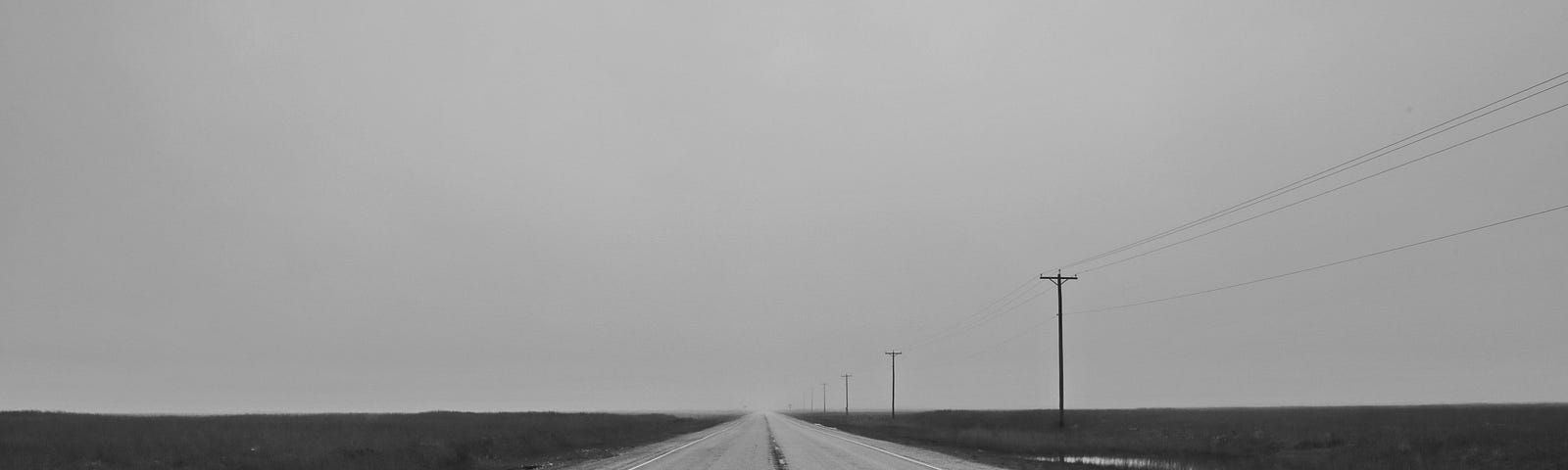 Black and white photo. Empty road stretching straight to horizon, through empty fields. No fences. Overcast day. Electric poles and wires stretched along right side of the road. A few shallow puddles.