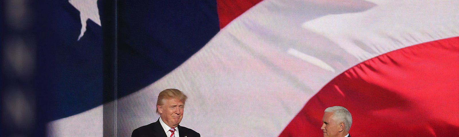 Background huge American flag, President Trump on left giving thumbs-up to Vice President Pence, on the right, who is smiling and applauding.