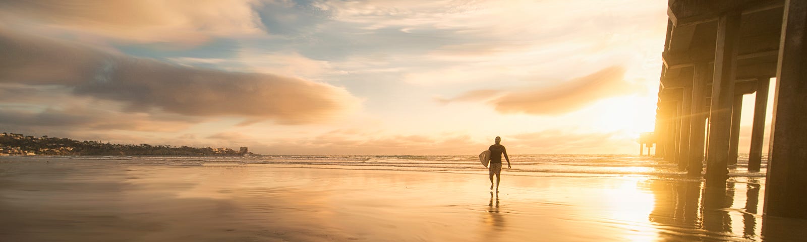 Man walking in wet sand toward sunset. Dock standing nearby.