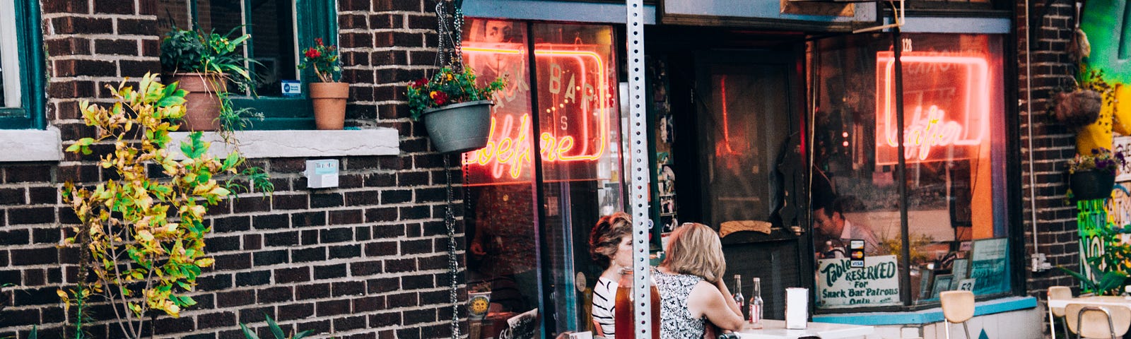 A bike is chained with a lamp post in fdront of a cafe with twinkling lights and some tables and chairs on the side of the footpath beside some potted plants.