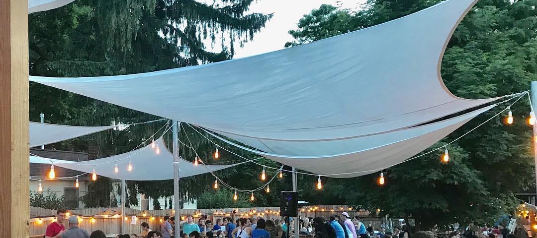 Cultivate Coffee & Tap House outdoor seating. Shade cloths and string lights hang above long tables full of community members having a great time drinking beer.