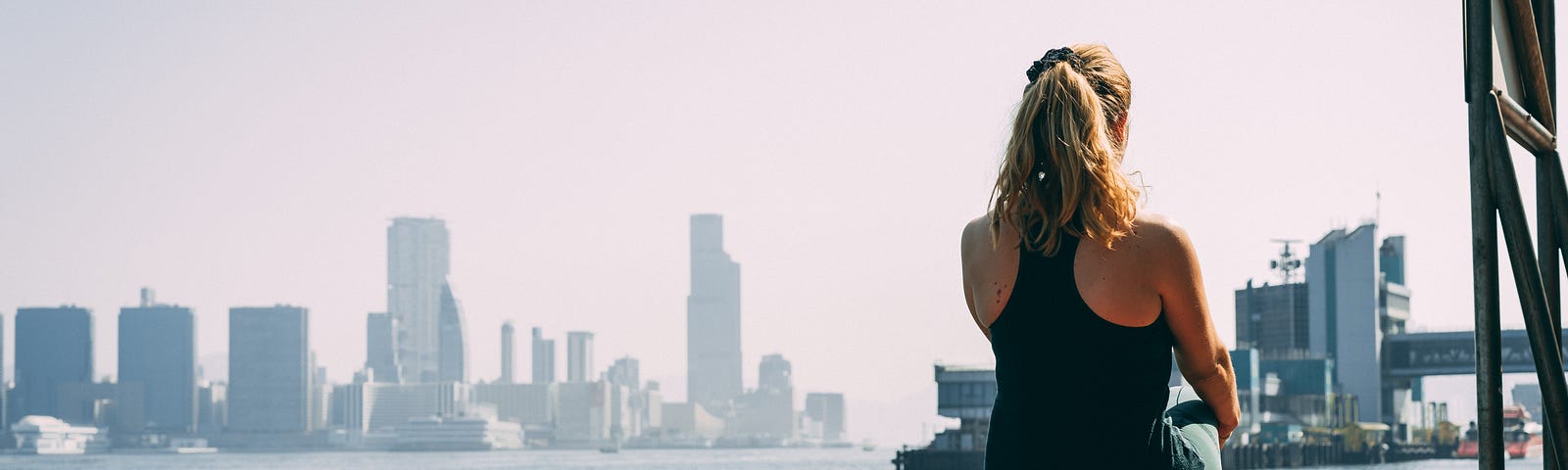 A blonde woman exercises while she looks out at the ocean. A city skyline is off in the distance