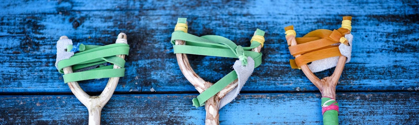 Three slingshots made from wood and rubber bands are laid out in a row on a blue painted wood table.