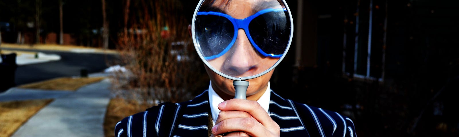 A man in a navy and white striped jacket, wearing blue framed sunglasses, holds a large magnifying glass, which reflects back a large and distorted view of his face.