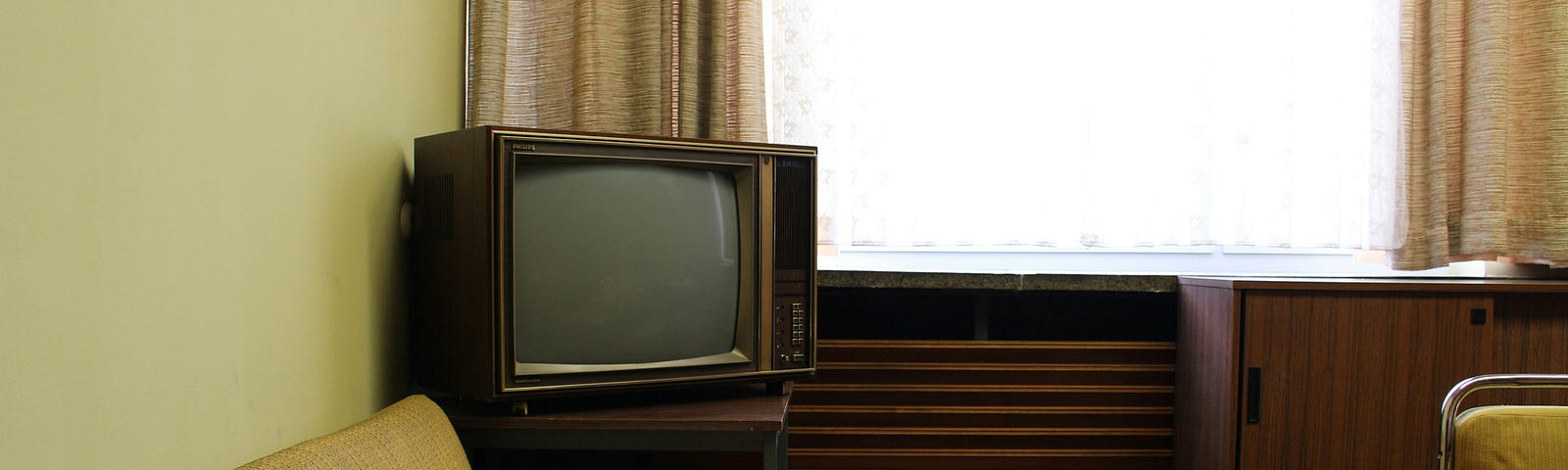 Vintage living room with a beige sofa, an old-fashioned television on a side table, and a wooden cabinet with a built-in record player, illuminated by natural light from a window.