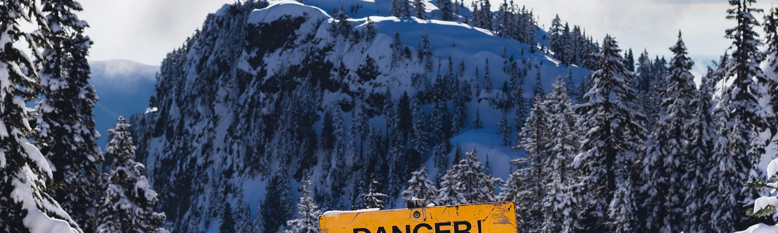 Sign warning of steep terrain and cliffs in front of snow-covered mountain and trees