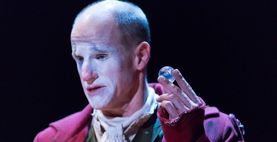 A performer in a red waistcoat and clown makeup holds up a large marble.