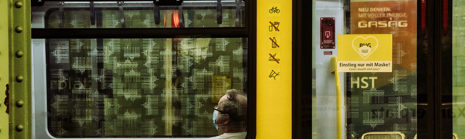 A yellow train from the outside with a sign showing that it is only allowed to enter with a mask. A man sits inside the train, wearing a mask.