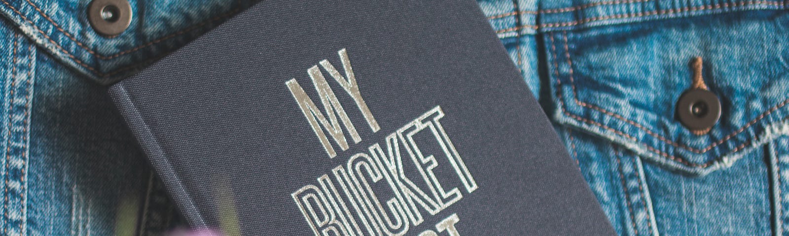 Image of a bucket list lying on a jean jacket with blurry flowers in the foreground.
