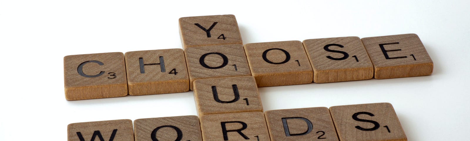Interlinked wooden letter tiles spell out the phrase “CHOOSE YOUR WORDS” on a white background