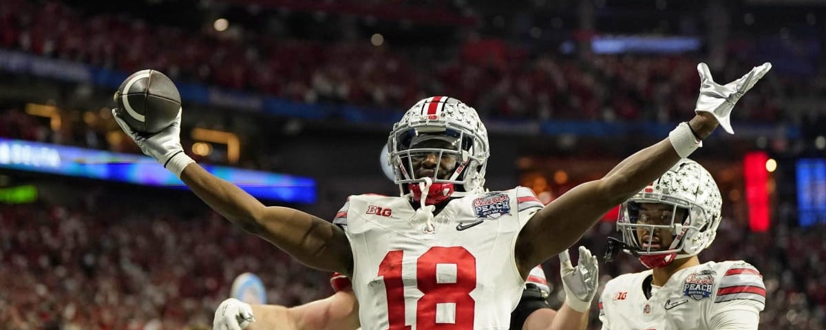 Ohio State wide receiver Marvin Harrison throws his two hands up in celebration, holding a football in his right hand.