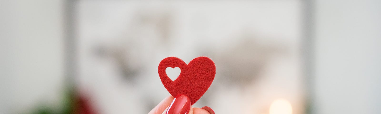 fFemale hand with red nail polish holding up a stiff red felt heart with a small heart-shaped hole in the upper left part. The background is a very blurry black and white map of the world with candles to the right and a plant to the left.