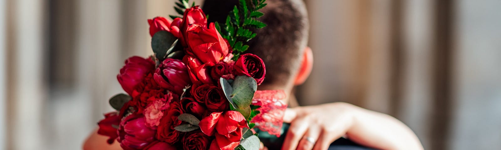 The back of a man is seen with someone’s arms around his shoulders, holding a red bouquet of flowers.