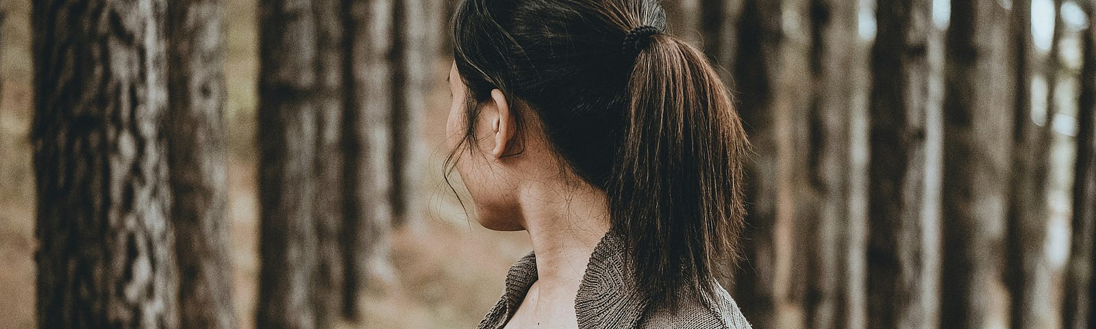 A brunette with a ponytail and an oversized cardigan is in the forest looking back at the trees.