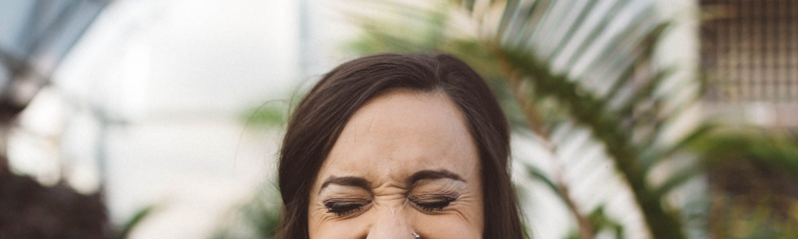 A woman with long brown hair, wearing a striped shirt and burgundy lipstick, is smiling and grimacing. Behind her are ferns and other plants.