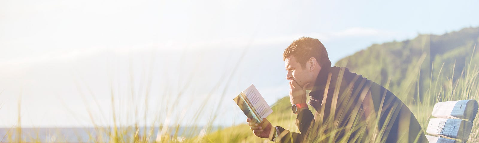 Man reading a book outdoors in a grassy field near a lake. He is sitting on an old bench.