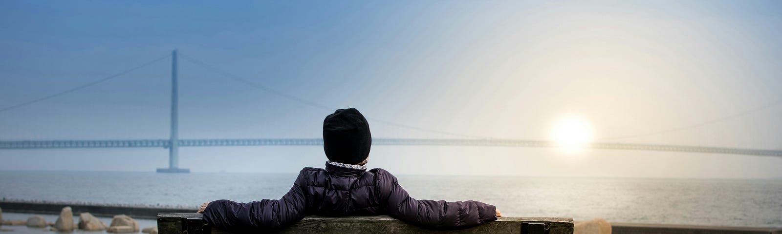 a man sitting on a park bench, overlooking a large bridge