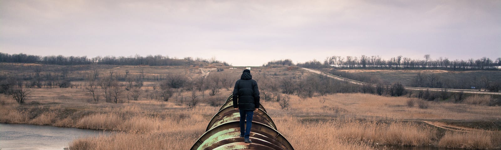 A jacketed man walks down the length of a rusty old pipeline.