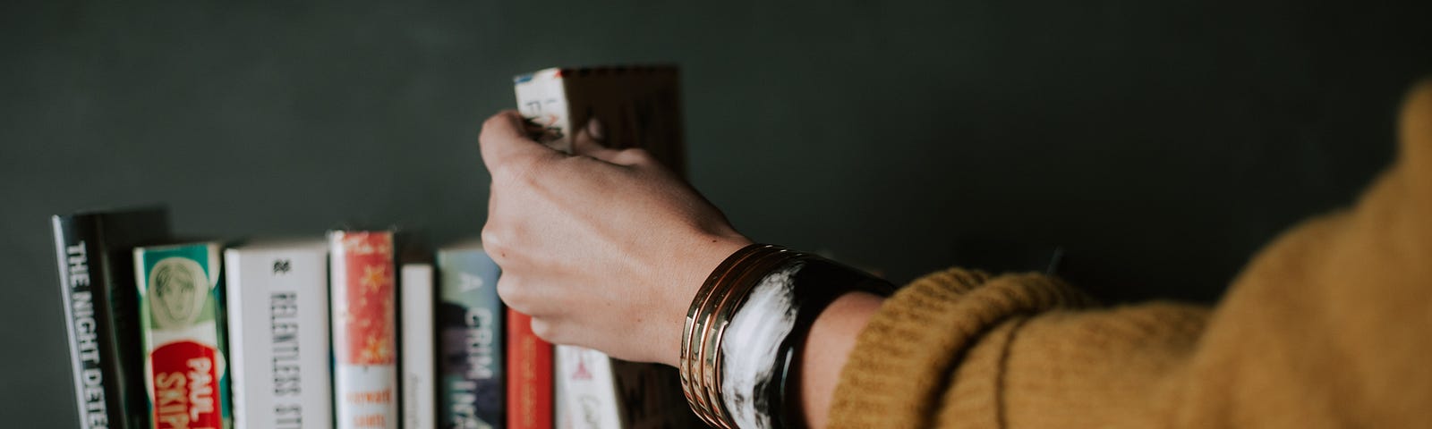 On a dark gray shelf sits a pile of various books. Someone’s hand can be seen picking up for placing a book onto the shelf.