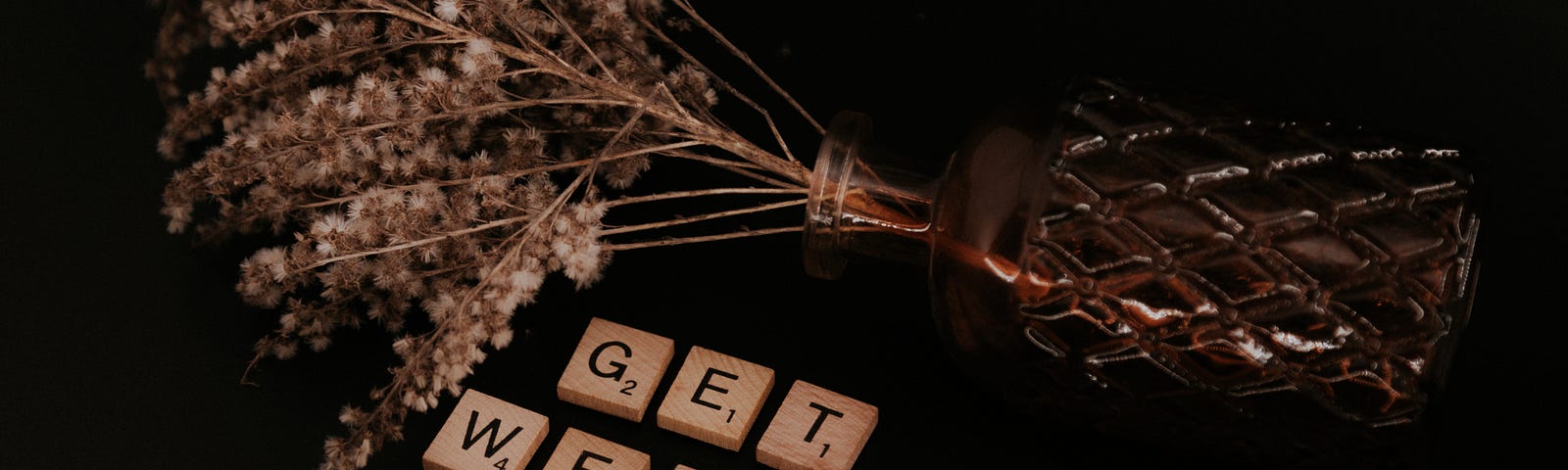 A bundle of dried flowers in a glass vase laying on its side. The words ‘Get Well Soon’ are laid out in Scrabble tiles beside it, on a black background.