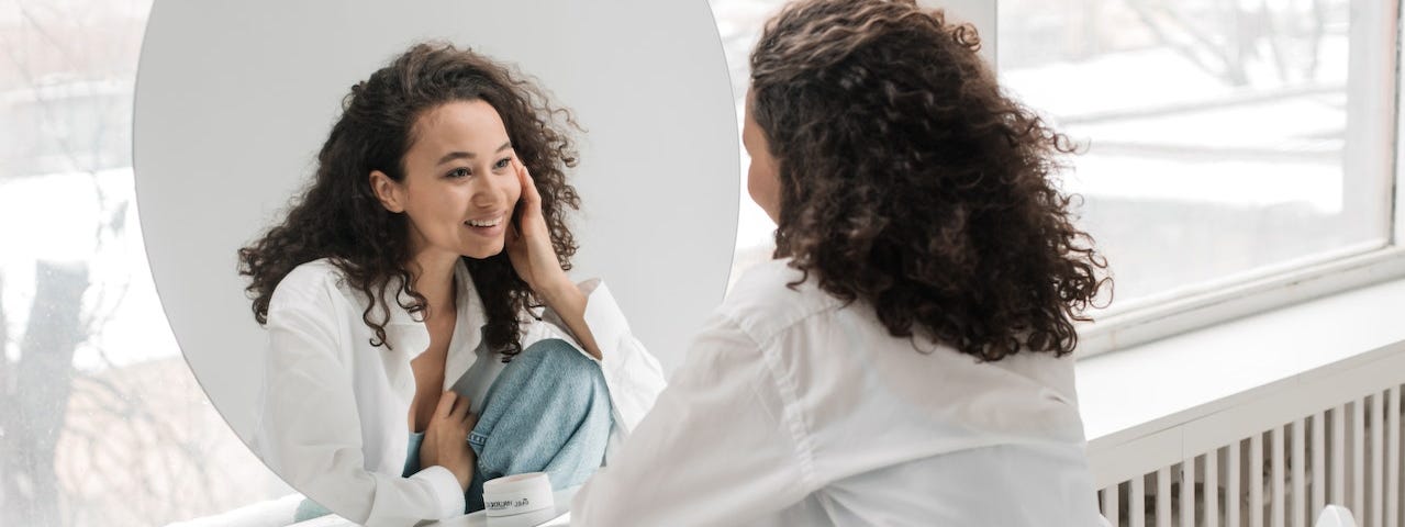 a woman in a white top smiles at herself in the mirror after doing her self-pleasure practice